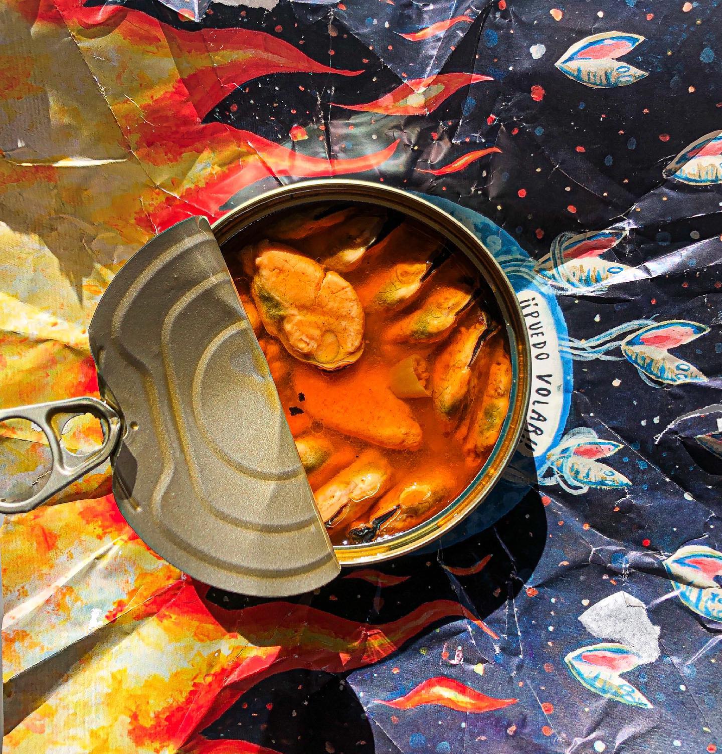 An overhead image of a tinned fish, featuring a tin of mussels resting on top of its artistic packaging. 