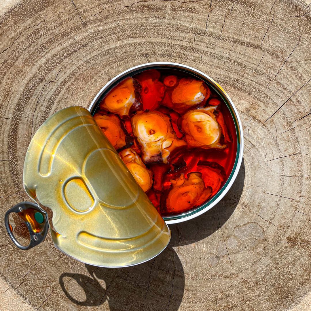 An open tin of La Curiosa Pulpo Gallego reveals tender pieces of marinated Galician octopus in vibrant red sauce, set against a rustic wooden background. The rich color and artisanal quality highlight traditional Spanish seafood preservation.