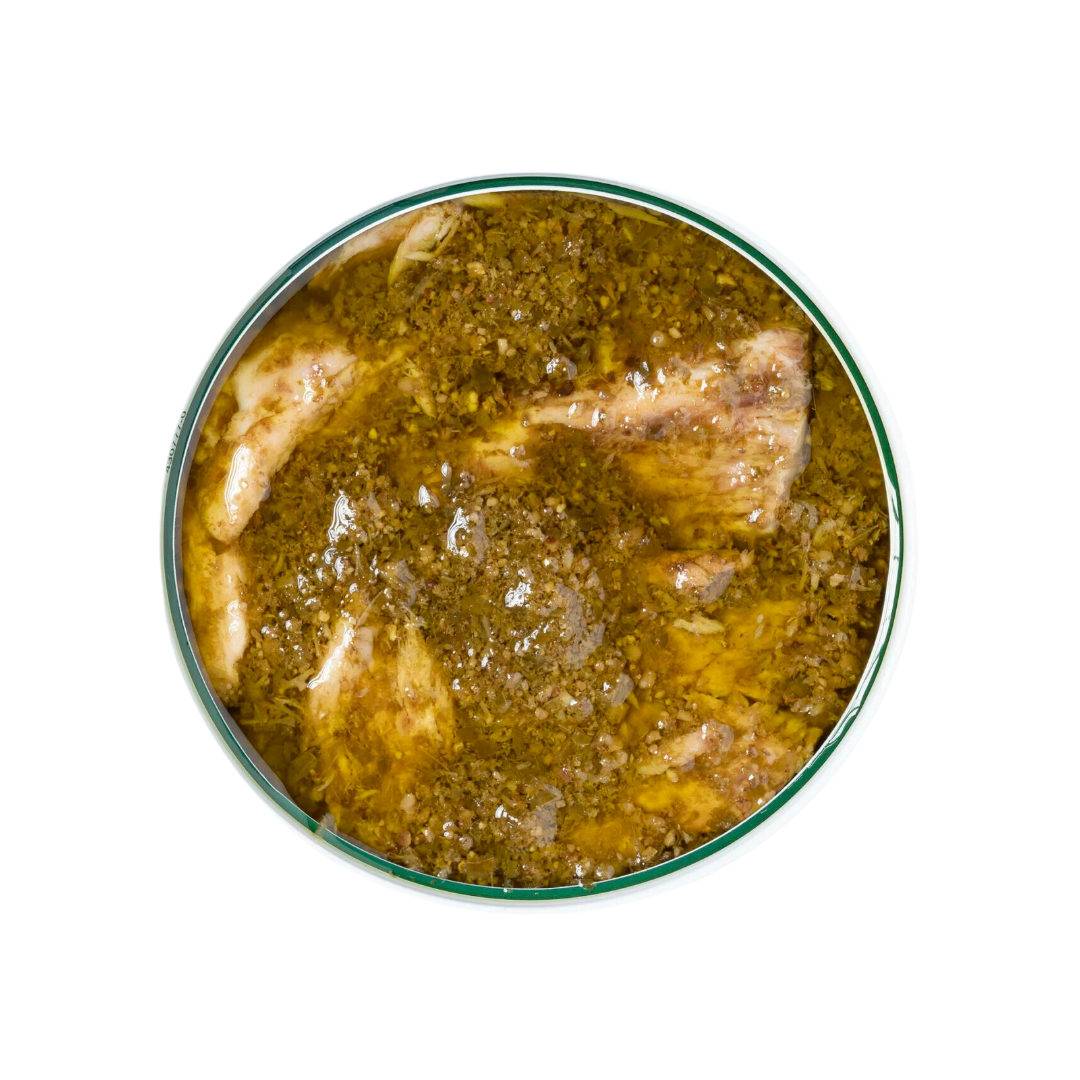 Top-down view of an open tin of yellowfin tuna belly in pesto. The tuna fillets are generously covered in a green, oily pesto sauce with visible herbs and seasoning. The tin is round with a green rim.