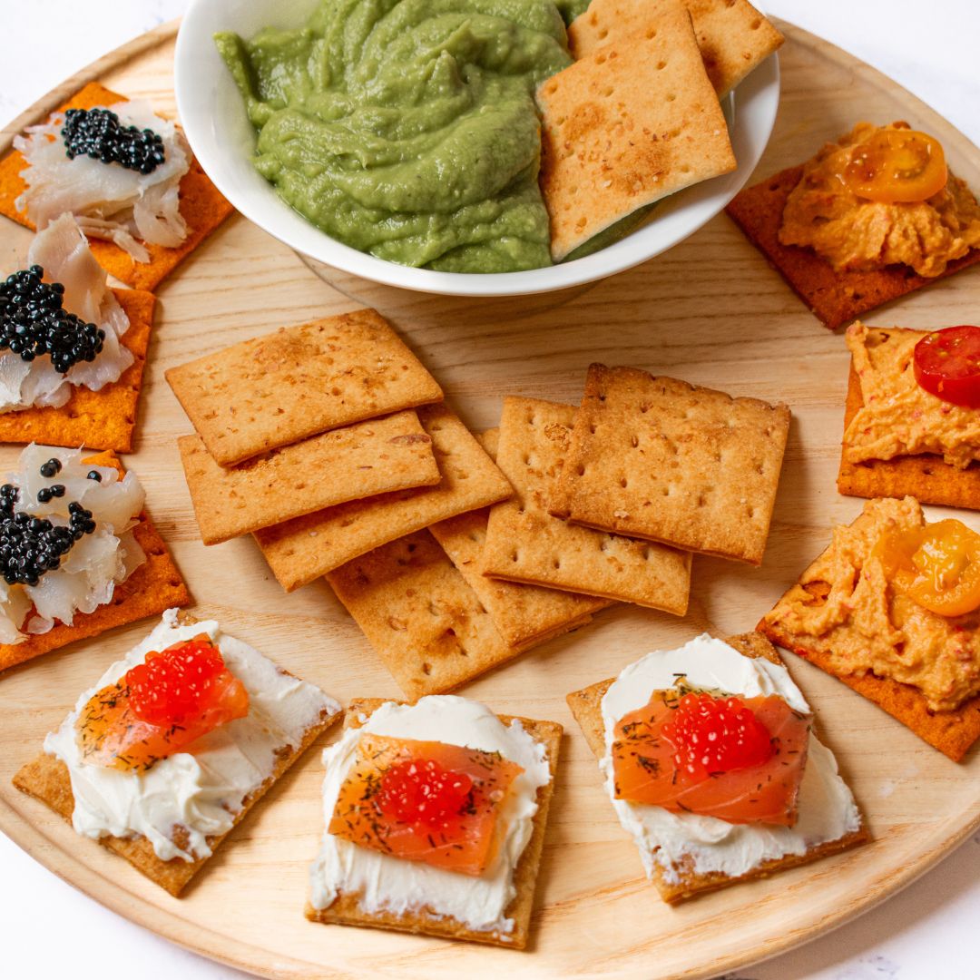 A round wooden serving board with a bowl of green dip in the center, surrounded by a variety of crackers. Some crackers are plain, while others are topped with spreads and garnishes: white fish with black caviar, creamy cheese with red caviar and dill, and a bright orange spread with halved cherry tomatoes.