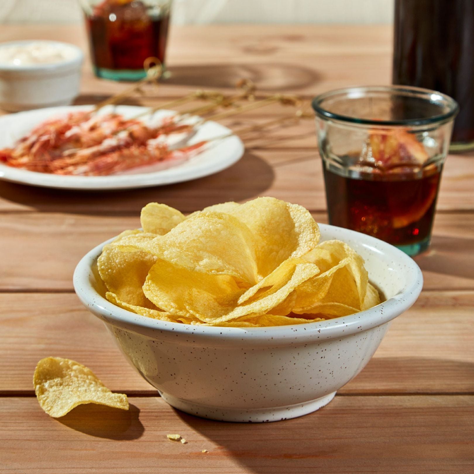 A rustic white bowl filled with golden Bonilla a la Vista potato chips sits front and center. In the background, red prawns on a plate, a ramekin of aioli, and glasses of vermut with citrus slices complete the laid-back tapas setup on a wooden table.