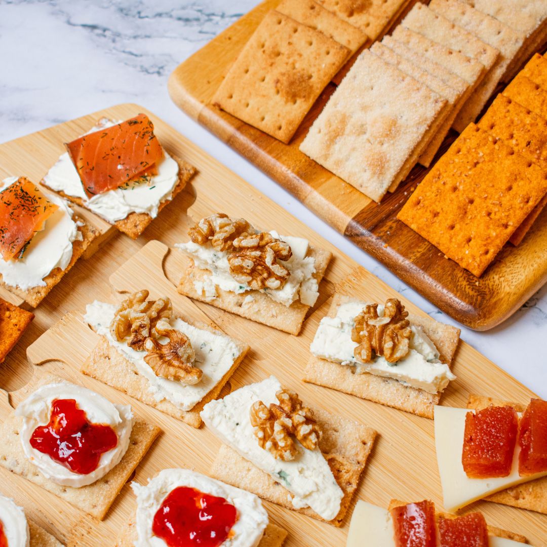 An assorted appetizer spread featuring various olive oil crackers topped with gourmet ingredients. The crackers are arranged on wooden boards and are topped with combinations such as cream cheese and smoked salmon, blue cheese with walnuts, cream cheese with red jam, and Manchego cheese with quince paste. In the background, there are plain and paprika-seasoned crackers neatly stacked. The setting has a light marble surface.