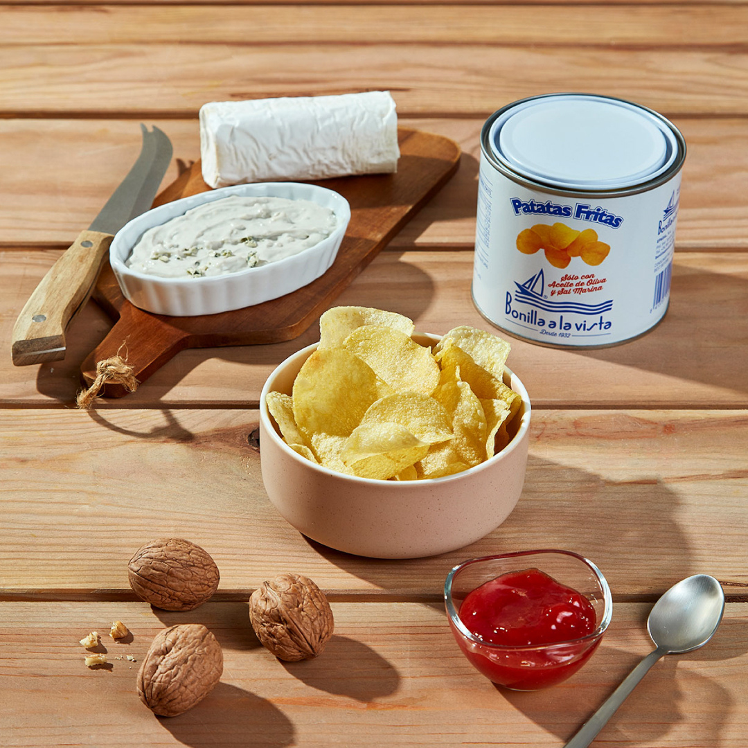 A 40g tin of Bonilla a la Vista potato chips sits next to a bowl of chips. Nearby, a goat cheese log, creamy blue cheese, walnuts, and a spoonful of red jam complete this sweet-and-savory tasting setup on a wooden table.