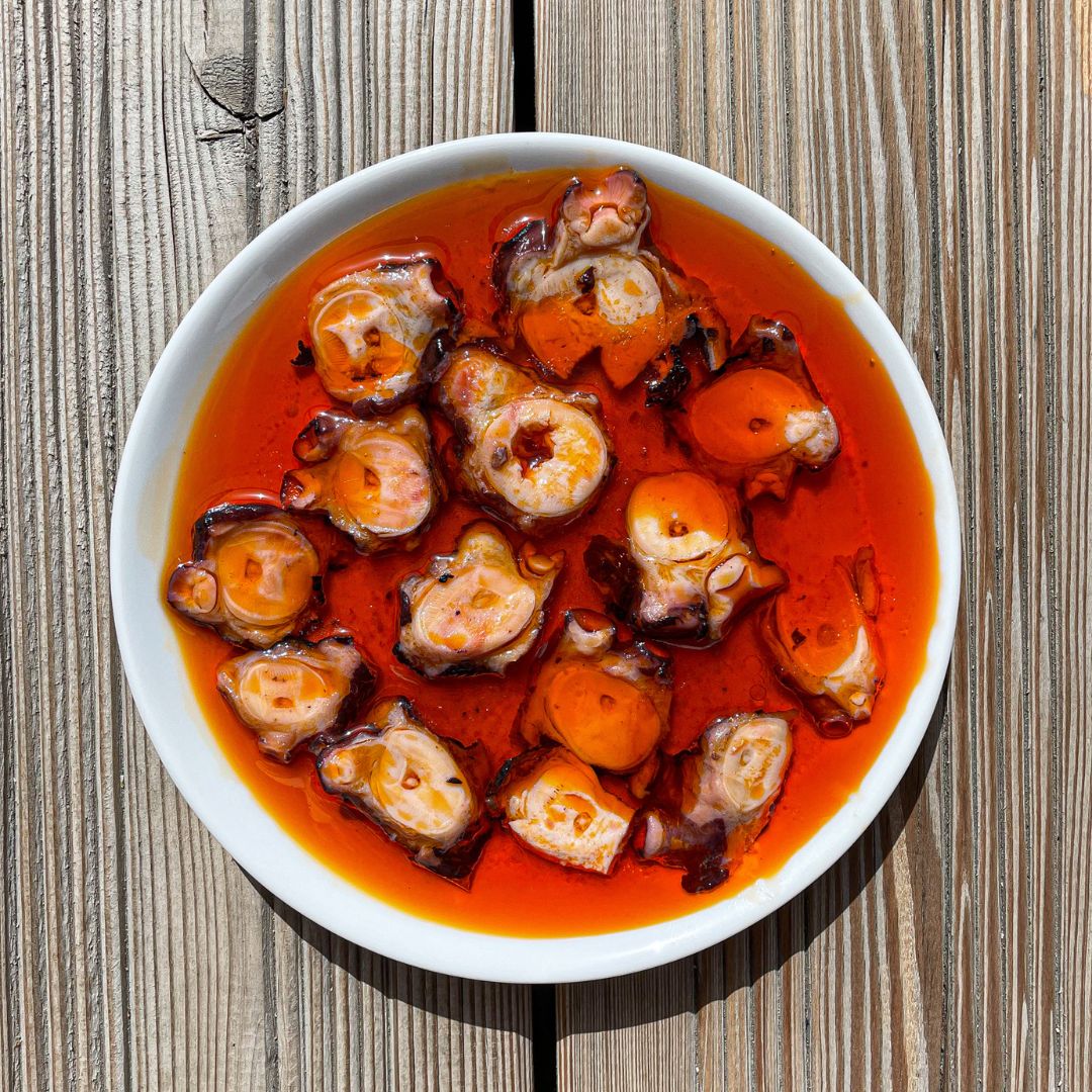 A white dish filled with vibrant slices of Galician octopus in rich red marinade, glistening under natural light on a rustic wooden table. The bold colors and artisanal cut evoke authentic Spanish seafood tradition.