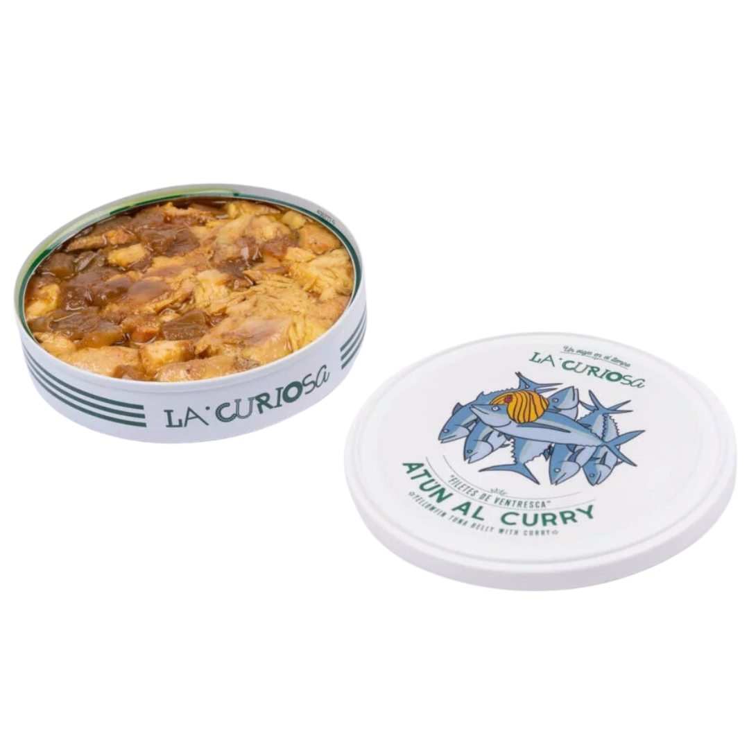 The open tin of La Curiosa Atún al Curry reveals tender yellowfin tuna belly pieces bathed in a vibrant, aromatic curry sauce. Chunks of onion glisten in the golden liquid, enhancing the dish’s rich texture and visual warmth. The nearby illustrated lid, playfully decorated with swimming tuna and a bold curry swirl, adds a touch of charm to this flavorful Spanish delicacy.