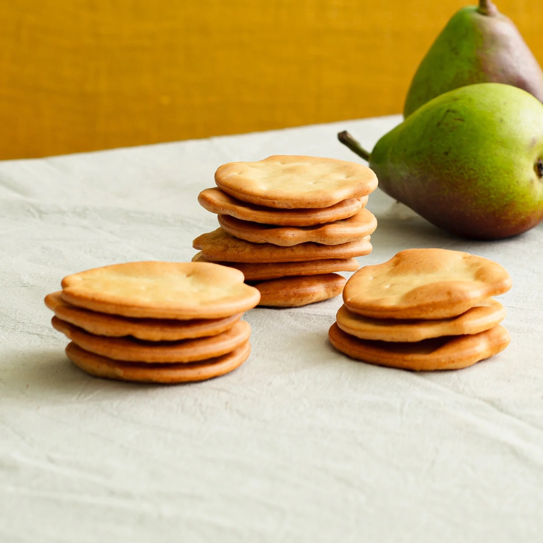 Stacks of golden-brown Spanish ship’s crackers sit on a white cloth with two green pears in the background. The crisp crackers feature a rustic, hand-formed look ideal for pairing with cheeses, tapas, or charcuterie.