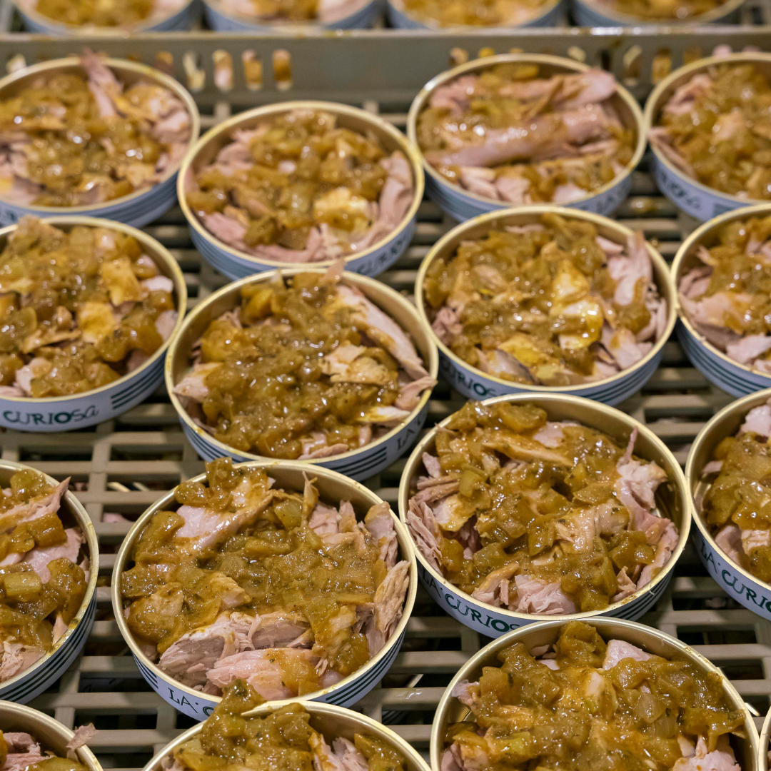 Dozens of tins of La Curiosa Atún al Curry are arranged in neat rows, each generously filled with succulent flakes of yellowfin tuna belly and topped with a golden curry-onion sauce. The vibrant mix of tender fish and aromatic seasoning glistens under the light, highlighting the artisanal care behind every tin in this small-batch Spanish production.