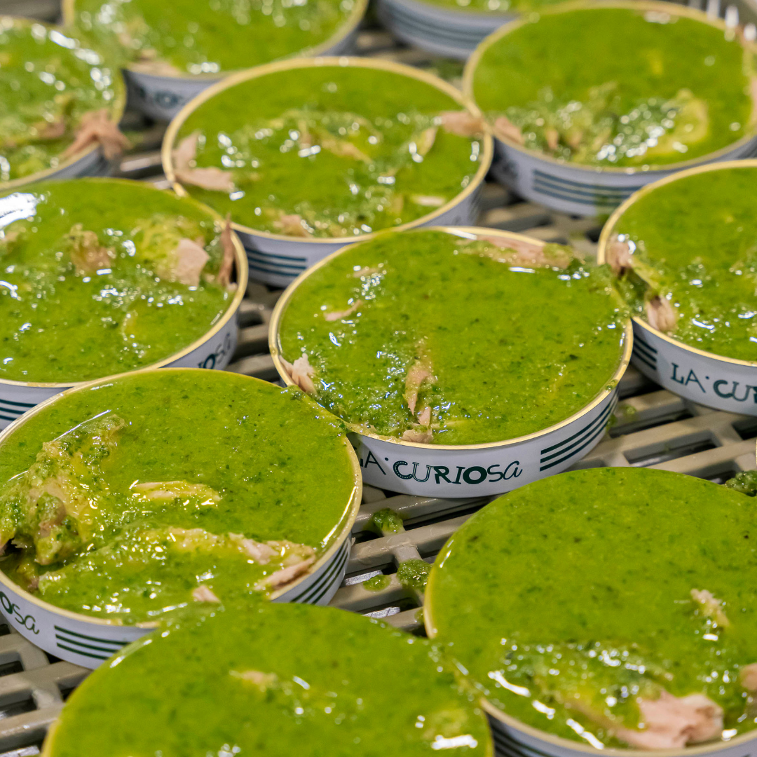 Close-up view of multiple tins of La Curiosa yellowfin tuna belly being filled with a bright green pesto sauce. The tins are neatly arranged on a production line tray, and the sauce visibly coats the tuna pieces inside each container, giving a fresh, vibrant appearance.