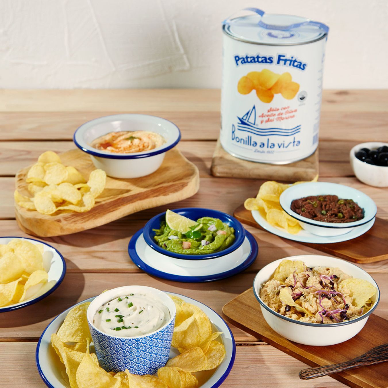 A large tin of Bonilla a la Vista potato chips sits at the back, unopened. Surrounding it are enamel bowls and plates of chips paired with colorful dips—creamy chive, guacamole, hummus and tuna salad. A bowl of black olives completes the vibrant, snack-ready spread on the wooden table.