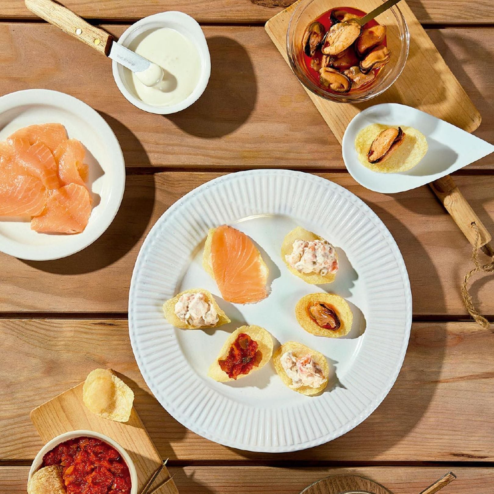 A white plate displays a selection of Bonilla a la Vista chips topped with smoked salmon, mussels, and tomato relish. Surrounding dishes offer extra toppings for DIY bites. A simple, elegant tapas presentation on a rustic wooden table.