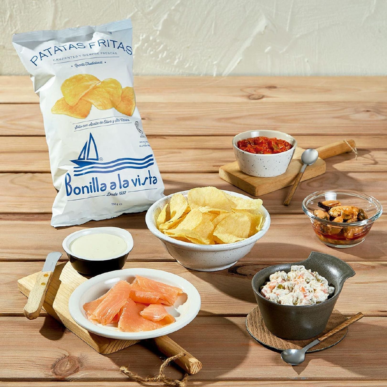 A 150g bag of Bonilla a la Vista potato chips leans casually on a wooden table. In front, a bowl of chips is paired with smoked salmon, creamy dressing, Russian salad, tomato relish, and a glass dish of mussels. A relaxed tapas spread with classic coastal flavors.