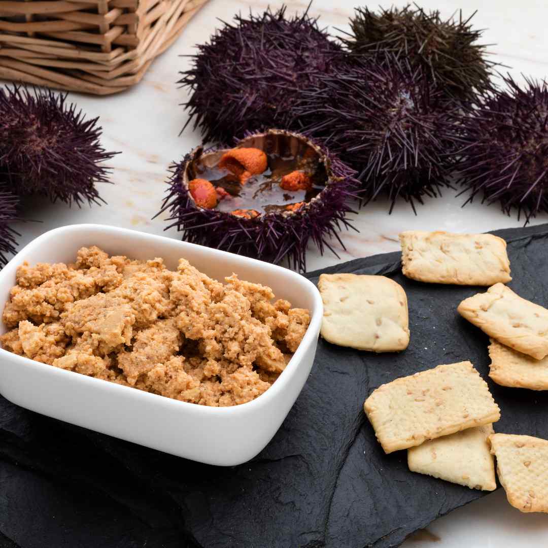 A close-up image showcasing a sea urchin pâté appetizer. In the foreground, a white bowl is filled with a crumbly, tan-colored sea urchin pâté placed on a black slate serving board. Beside the bowl, there are several pieces of square, golden crackers. In the background, unopened and one opened sea urchin shell with bright orange roe are displayed, with a wicker basket partially visible. The presentation suggests a gourmet seafood snack or appetizer setting.