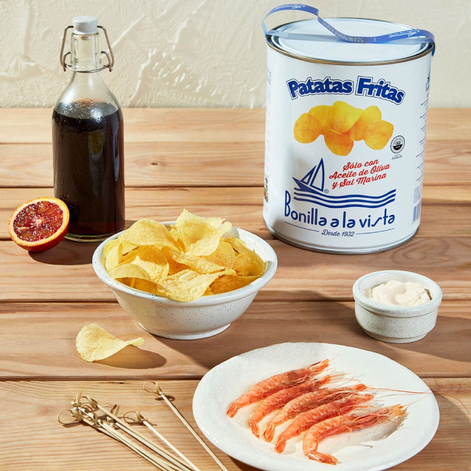 A large tin of Bonilla a la Vista potato chips with a sailboat graphic and bold “Patatas Fritas” text. In front, a white bowl filled with golden chips. Surrounding it are red prawns on a plate, a small pot of aioli, a glass bottle of vinegar, and half a blood orange. Wooden skewers rest casually nearby.