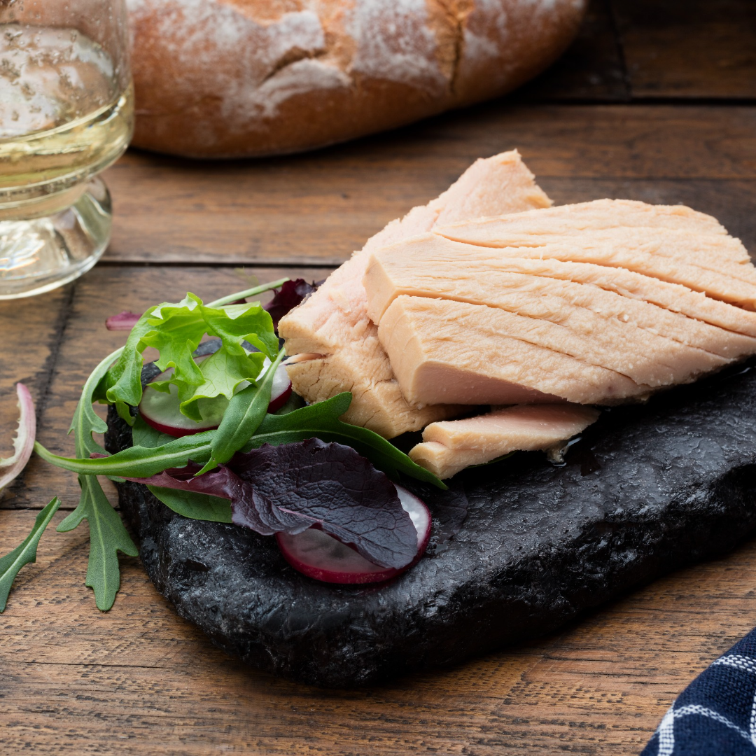 Thick, flaky fillets of white albacore tuna are artfully arranged on a rustic black slate, paired with a medley of fresh greens and sliced radish. A crusty bread loaf and a glass of white wine complete this elegant Spanish seafood presentation.