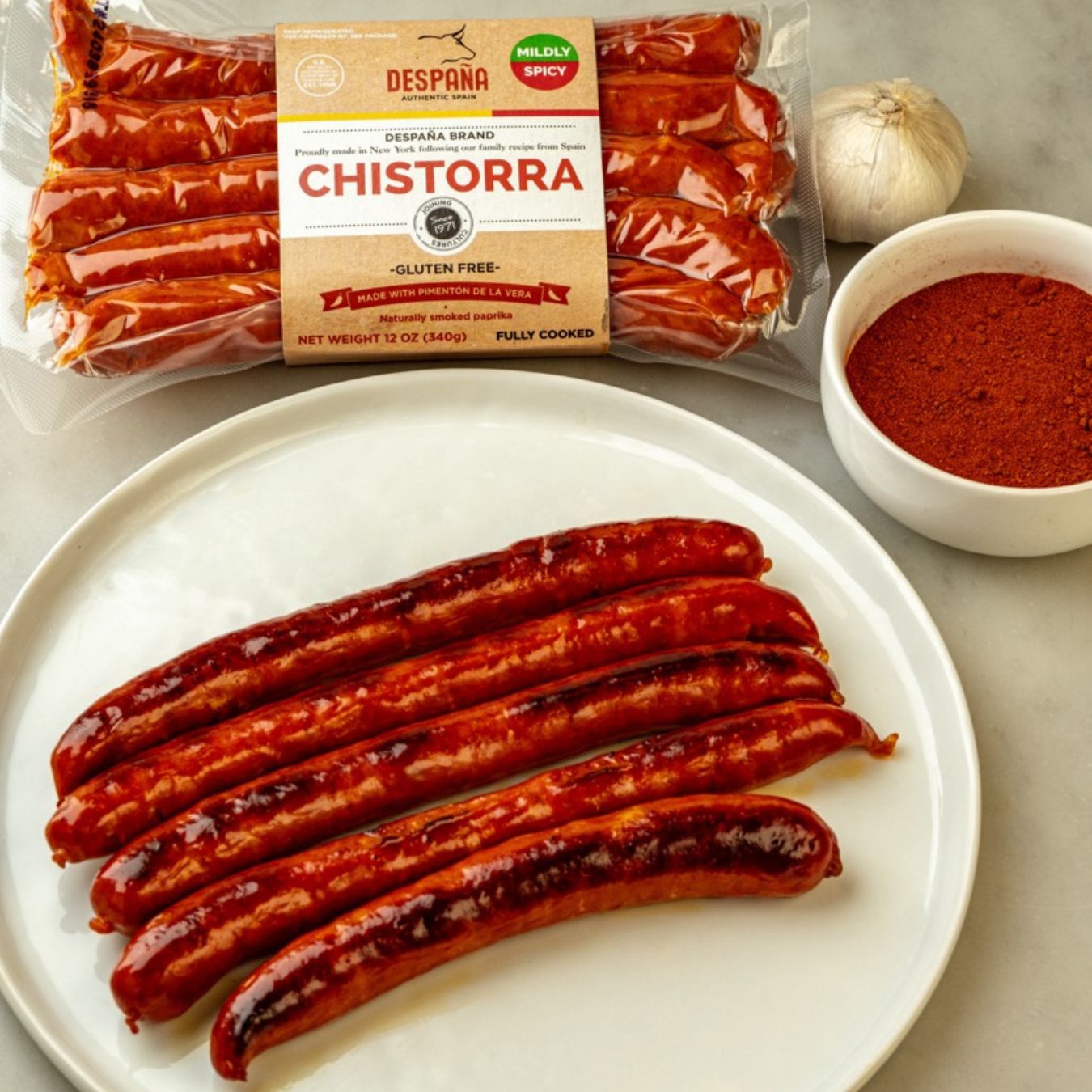 A pack of Despaña Chistorra sits above a plate of glistening, cooked sausages. Beside them, a bulb of garlic and a bowl of smoked paprika hint at the bold, traditional Spanish flavors inside.
