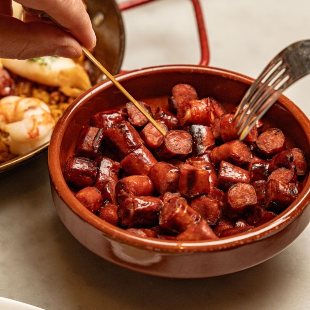 A rustic terracotta dish filled with golden-browned, bite-sized chistorra pieces. A skewer and fork reach in, ready to snack. In the background, a glimpse of seafood paella adds to the rich tapas atmosphere.