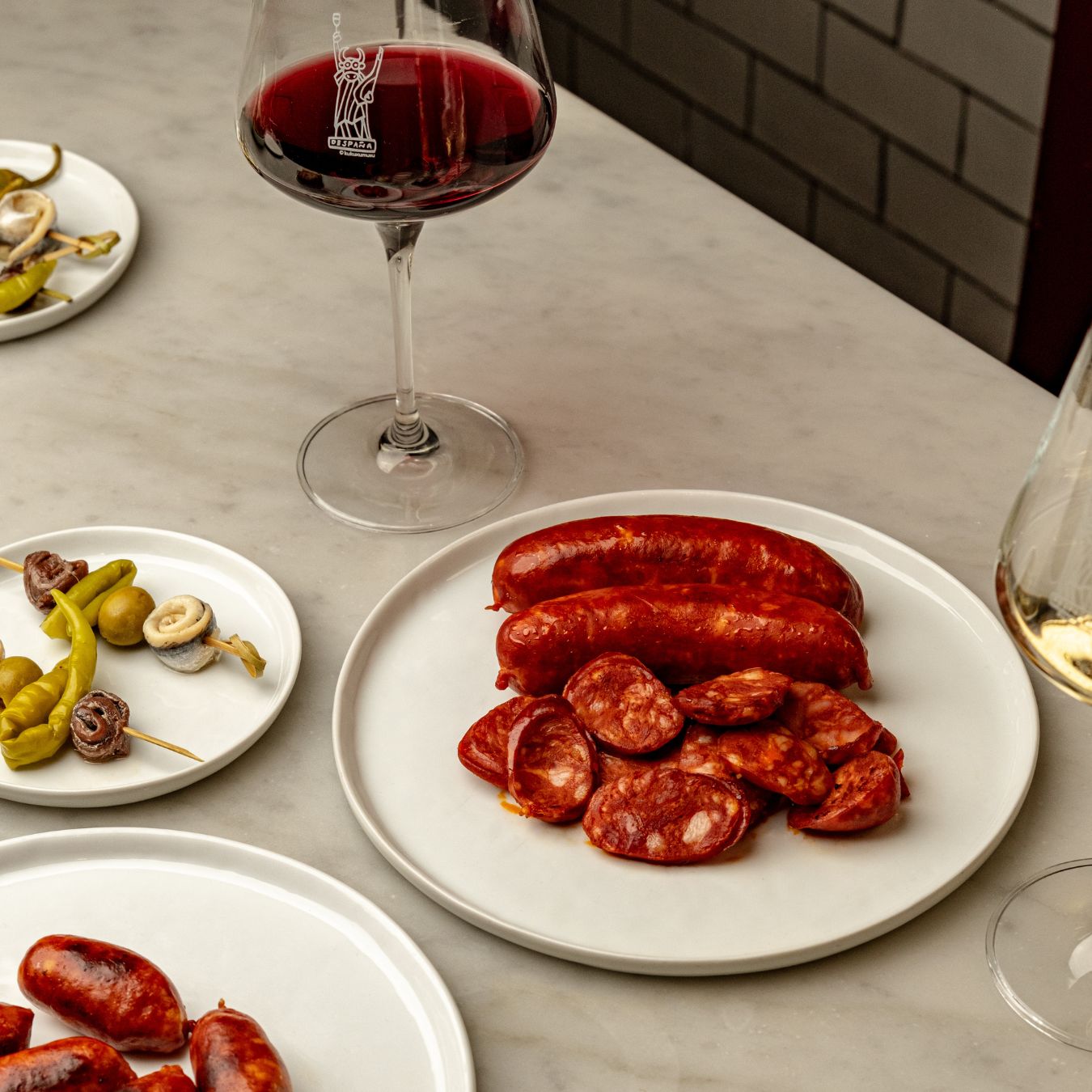 A plate of sliced and whole Spanish chorizo served alongside pickled vegetables and wine on a marble table. The rich red sausage, seasoned with smoked paprika, pairs elegantly with glasses of red and white wine, creating a classic tapas-style setting.