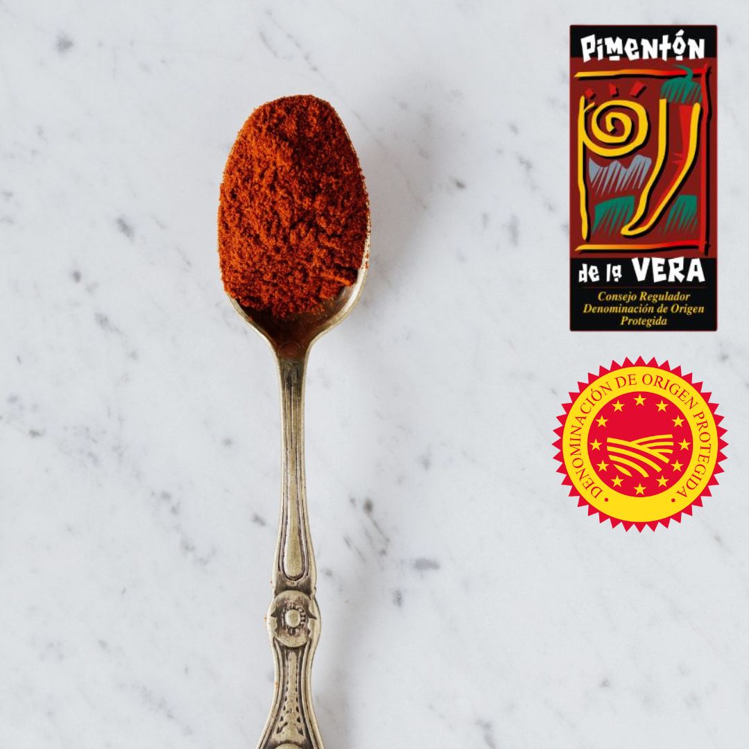 A spoonful of vibrant Pimentón de la Vera Dulce rests on white marble, showcasing its fine texture and deep red hue. Two DOP certification badges highlight its authenticity and origin from La Vera, Spain. This sweet smoked paprika is essential for adding bold, smoky flavor to traditional Spanish cuisine.