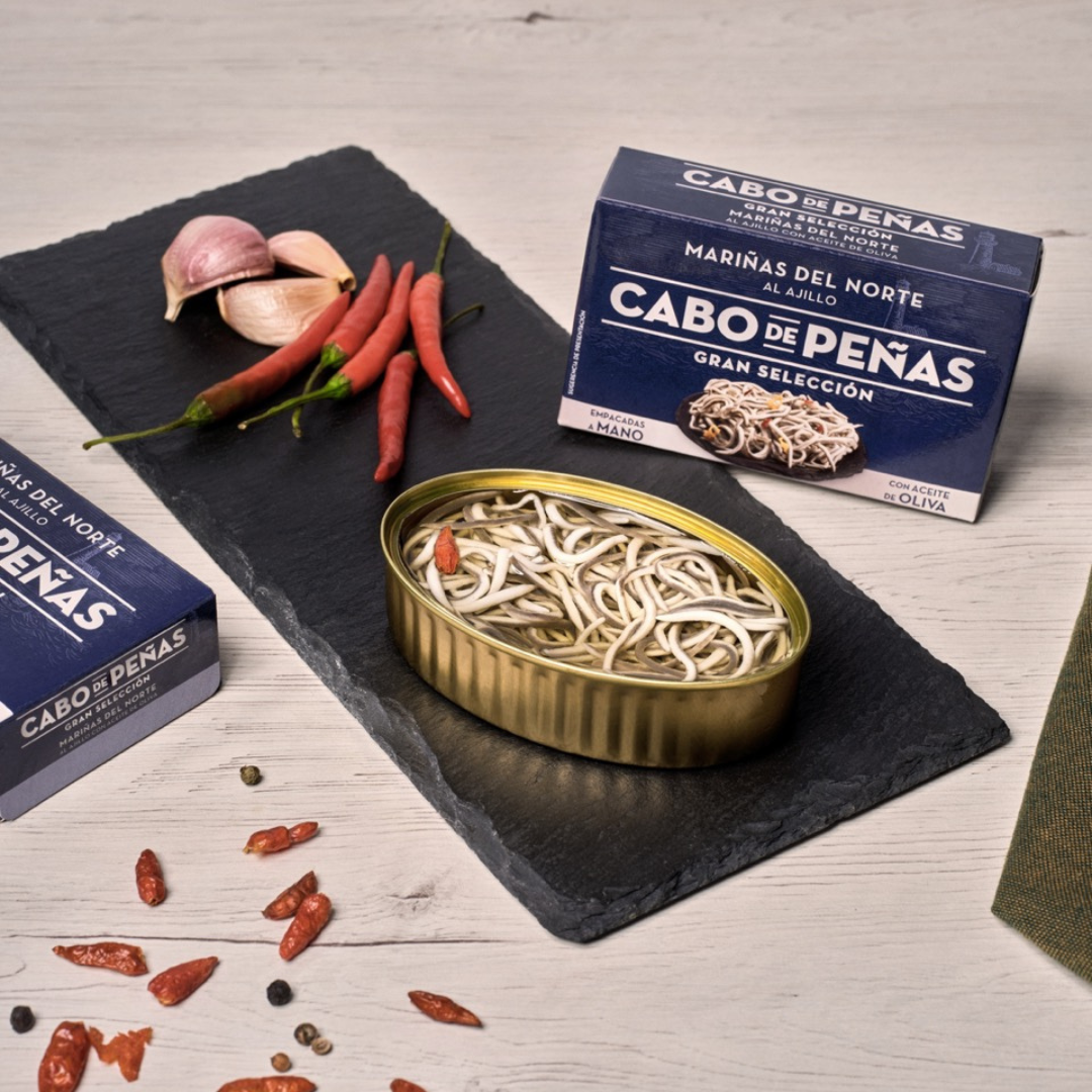 Open tin of Cabo de Peñas Surimi Baby Eel in spicy olive oil on a black slate with whole red chili peppers and garlic cloves. The packaging and ingredients emphasize traditional Spanish flavors and artisanal seafood craftsmanship.