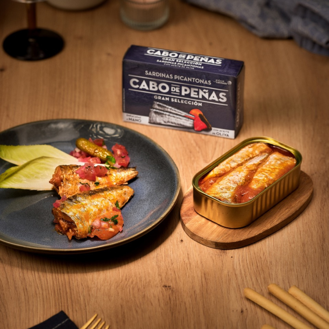 A serving of Cabo de Peñas Gran Selección spicy sardines is presented on a dark ceramic plate with tomato garnish, pickles, and endive leaves. Beside the plate, an open tin of sardines in spiced olive oil sauce is displayed on a small wooden board. In the background, a branded box of Cabo de Peñas sardines is visible, indicating the product name "Sardinas Picantonas" and emphasizing the use of olive oil and natural ingredients.