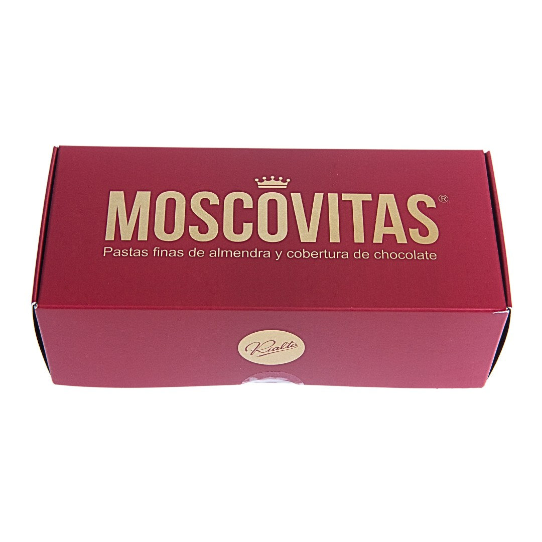 Angled view of a red rectangular box labeled "MOSCOVITAS" in large gold lettering, with a crown icon above the text. Below, it reads “Pastas finas de almendra y cobertura de chocolate.” The side of the box features a geometric pattern in gold, white, and red, with the brand name "Rialto" centered in a circular emblem.