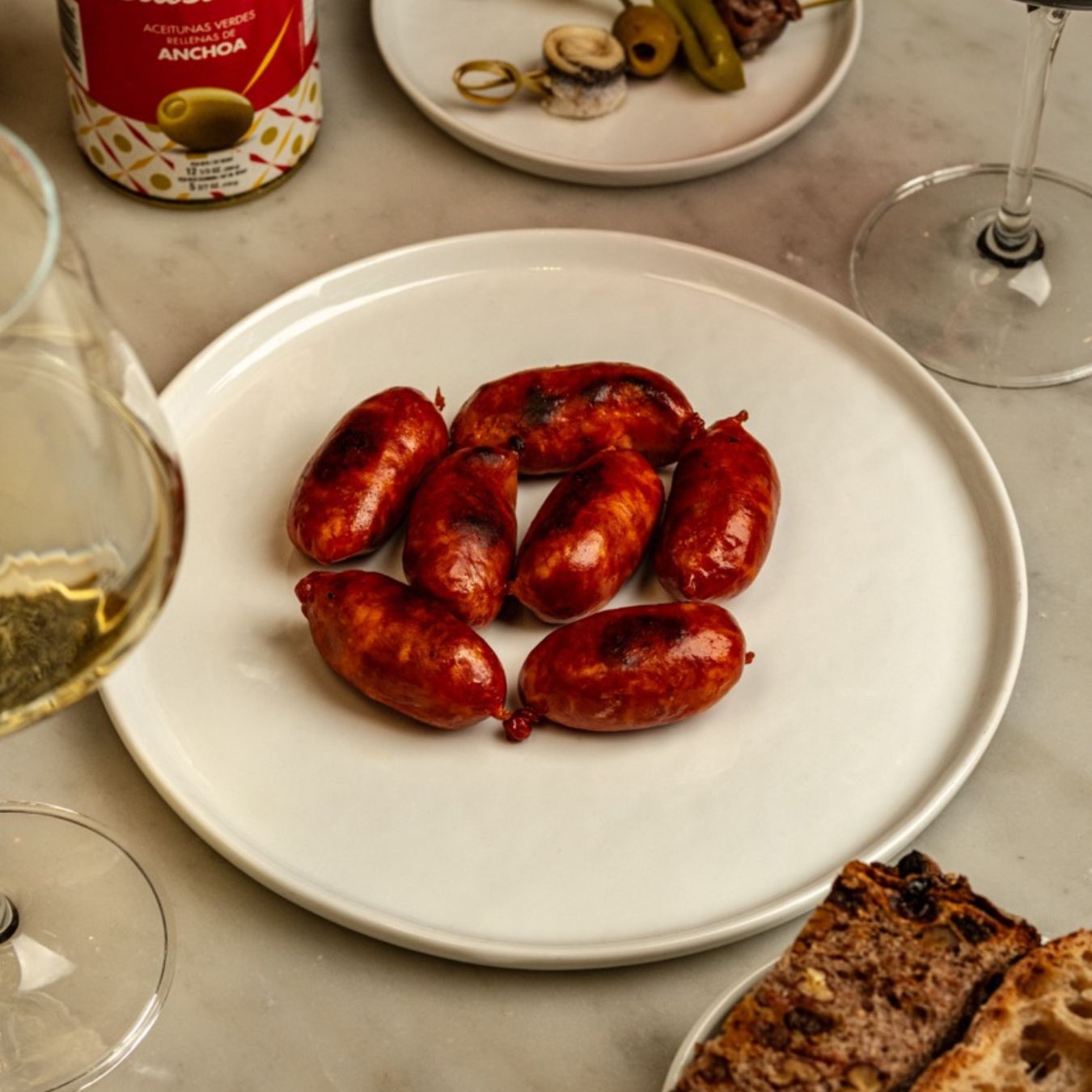 A white plate of seared mini chorizos sits on a marble table, their glossy, charred skins glistening. Surrounding the dish are wine glasses, anchovy-stuffed olives, and rustic bread—classic Spanish tapas elegance.