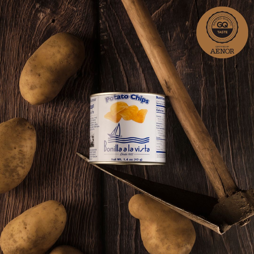 A 40g tin of Bonilla a la Vista potato chips displayed on dark wood among raw potatoes and a rustic digging tool. The GQ Taste award badge highlights its gourmet quality, tying the farm-to-snack story together.