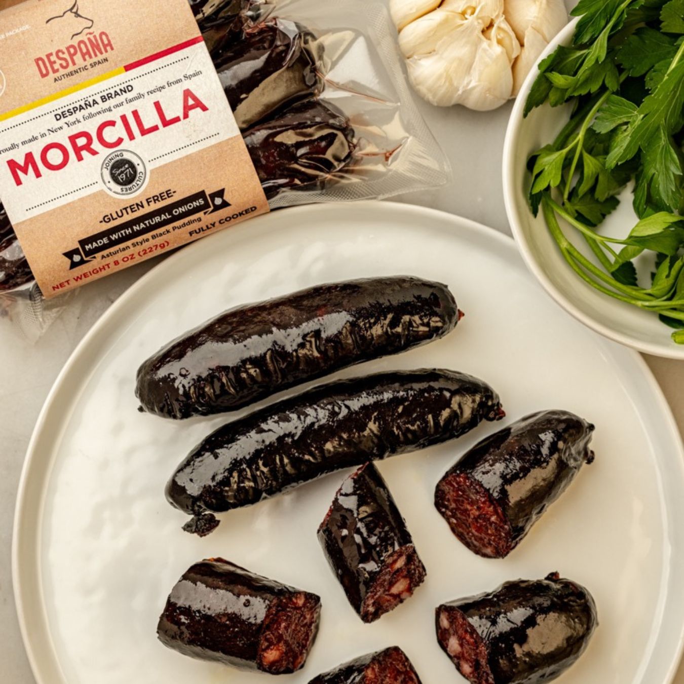 Despaña Morcilla sausages, whole and sliced, arranged on a white plate. Their glossy black casing reveals a rich, dark filling. In the background, the vacuum-packed label is visible alongside fresh parsley and garlic.