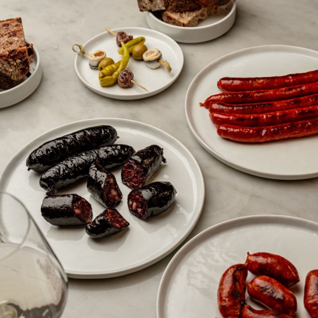 An elegant marble table spread with sliced morcilla, chistorras, mini chorizos, and gildas. Rustic bread and a glass of white wine complete this rich Spanish charcuterie scene.