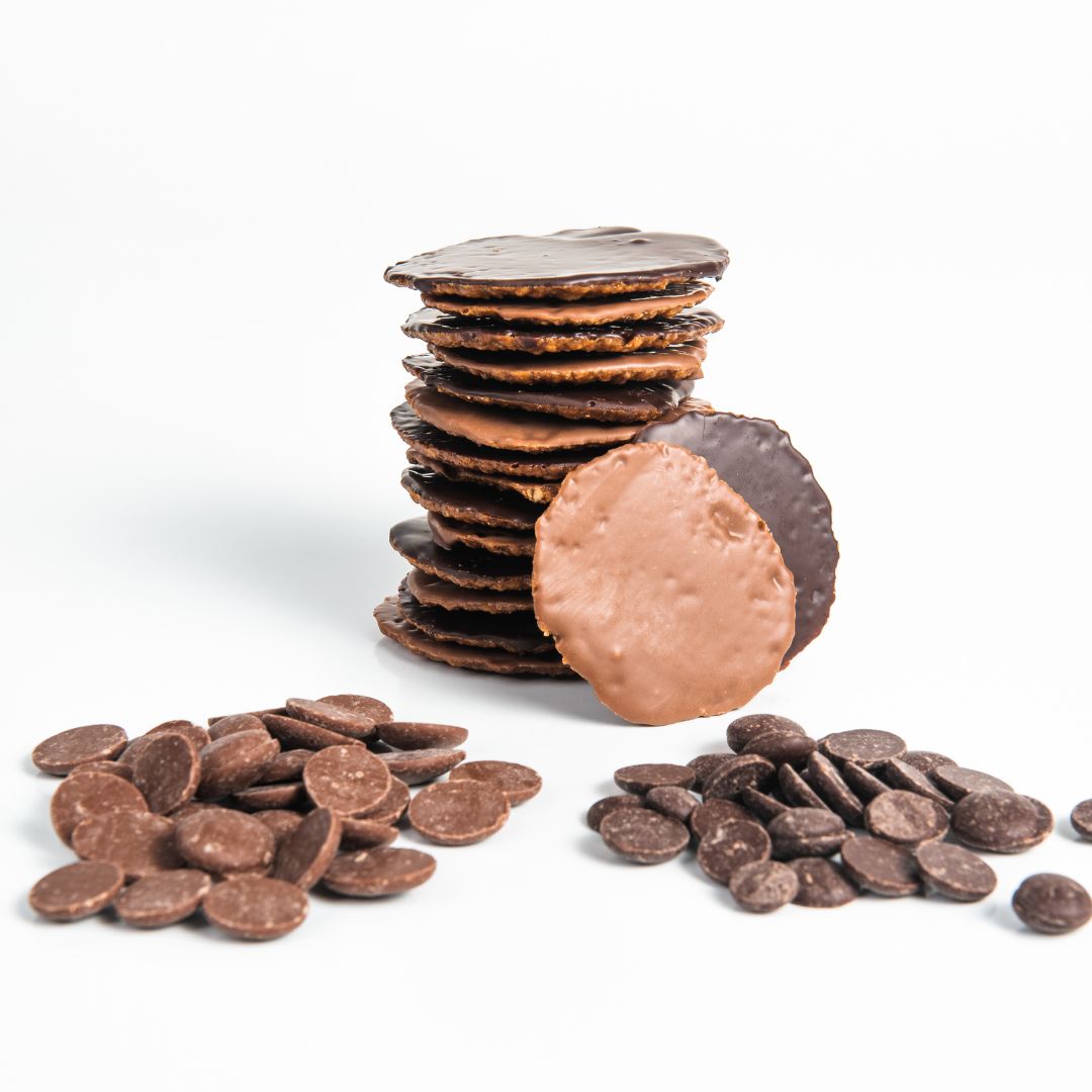 A stack of round Moscovitas cookies coated in dark and milk chocolate, with two cookies leaning in front to display both chocolate types. Surrounding the cookies are scattered chocolate discs used for coating. The background is white and minimalistic.