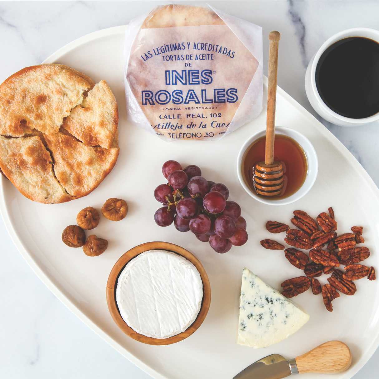 A rustic serving tray displays Ines Rosales Sweet Olive Oil Tortas surrounded by gourmet pairings—figs, red grapes, pecans, and creamy cheeses. A drizzle of honey and a cup of black coffee complete this elegant Spanish-inspired breakfast or snack spread.