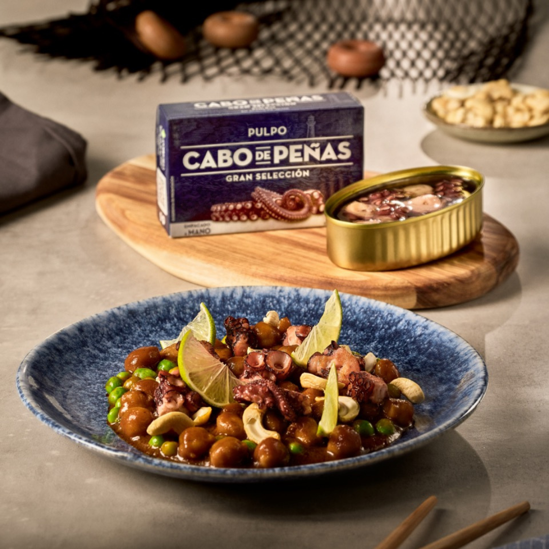A plate that has been filled beautifully with chickpeas and tinned Cabo de Peñas octopus. It has been garnished with peas and lime.