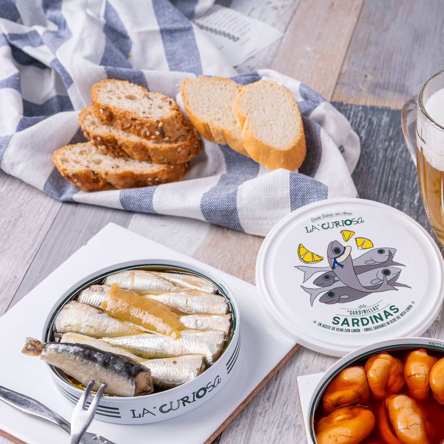 An open tin of La Curiosa sardines in olive oil with lemon takes center stage, its contents shimmering beside plump butter beans and a frothy glass of beer. Rustic slices of seeded and white bread rest atop a casually draped checkered cloth, setting the scene for a relaxed, flavor-rich tapa moment. The illustrated lid featuring lemon-adorned sardines adds whimsy to this inviting Spanish spread.
