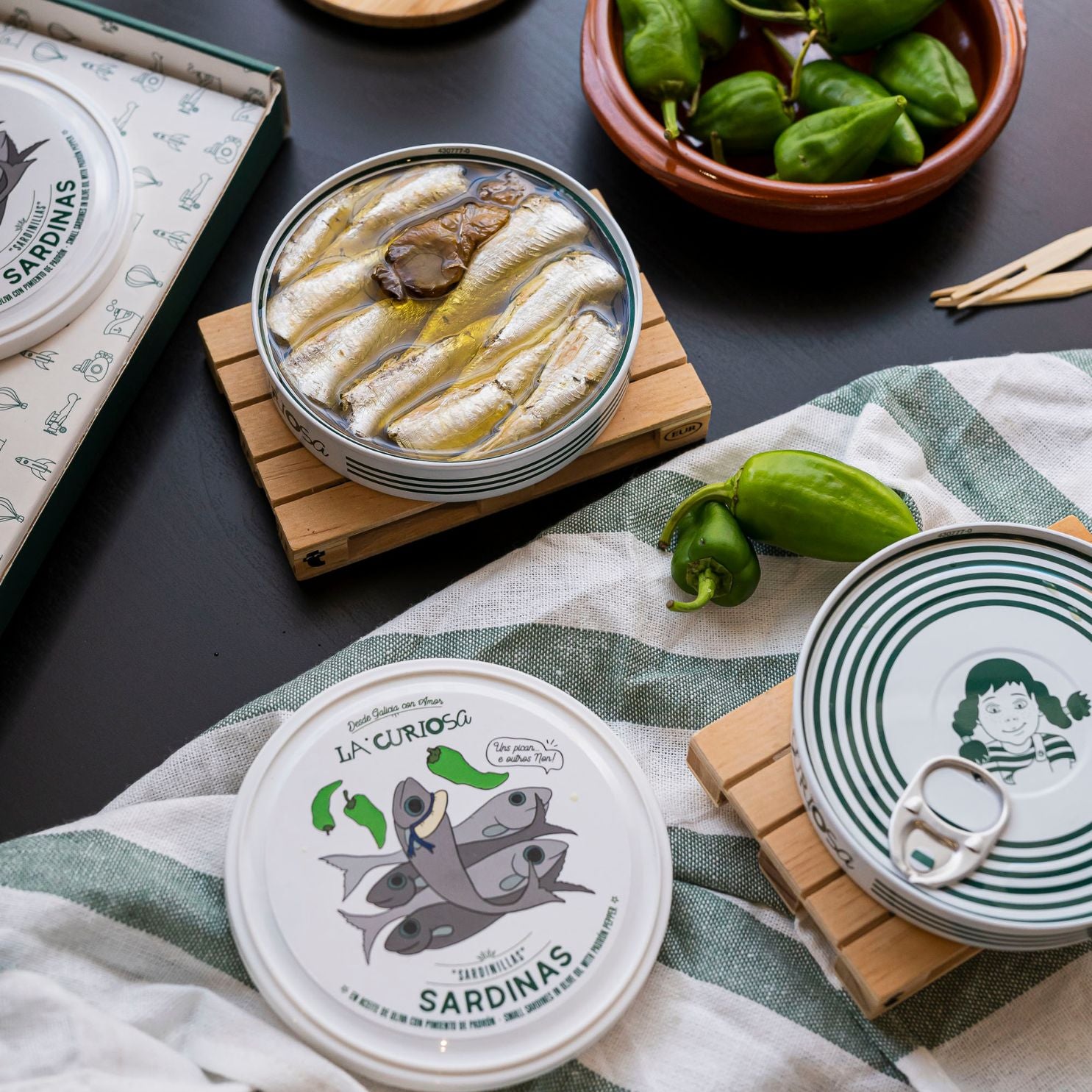 An open tin of La Curiosa sardines rests on a wooden coaster, revealing glistening fish and padrón peppers in golden olive oil. Nearby, fresh peppers and closed tins with playful designs evoke a gourmet yet whimsical Galician snacking scene.