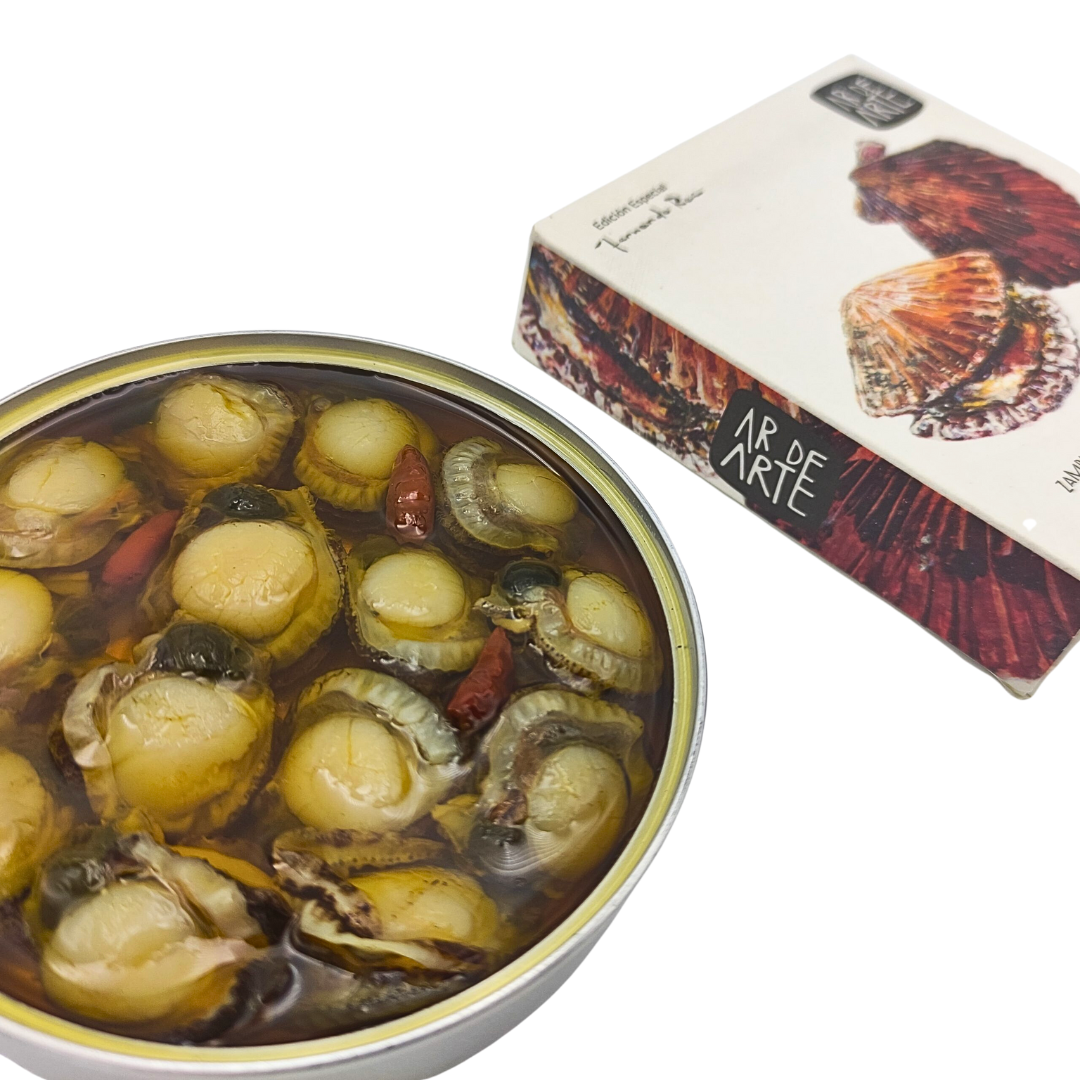An open tin of Ar de Arte Galician scallops in olive oil with garlic and chili, showcasing tender, bite-sized scallops with whole chilies in golden oil. The artisanal box in the background features vivid shell illustrations, reflecting the premium Spanish seafood inside.