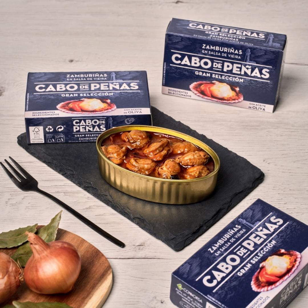An open tin of Cabo de Peñas zamburiñas in vieira sauce rests on a slate board, surrounded by product boxes and fresh shallots. The tender small scallops are coated in rich, red Galician sauce made with olive oil, highlighting traditional Spanish seafood excellence.