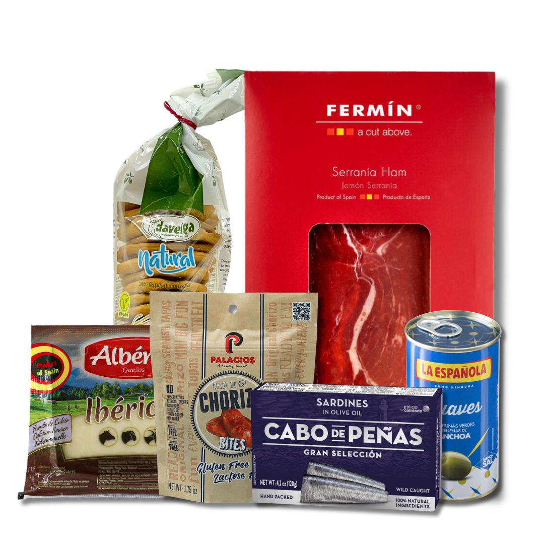 A collection of Spanish snacks displayed in front of a red gift box labeled 'Fermin'. The items include pre-sliced jamon, chorizo bites, iberico cheese, olives with anchovy, sardines in olive oil, and olive oil crackers.