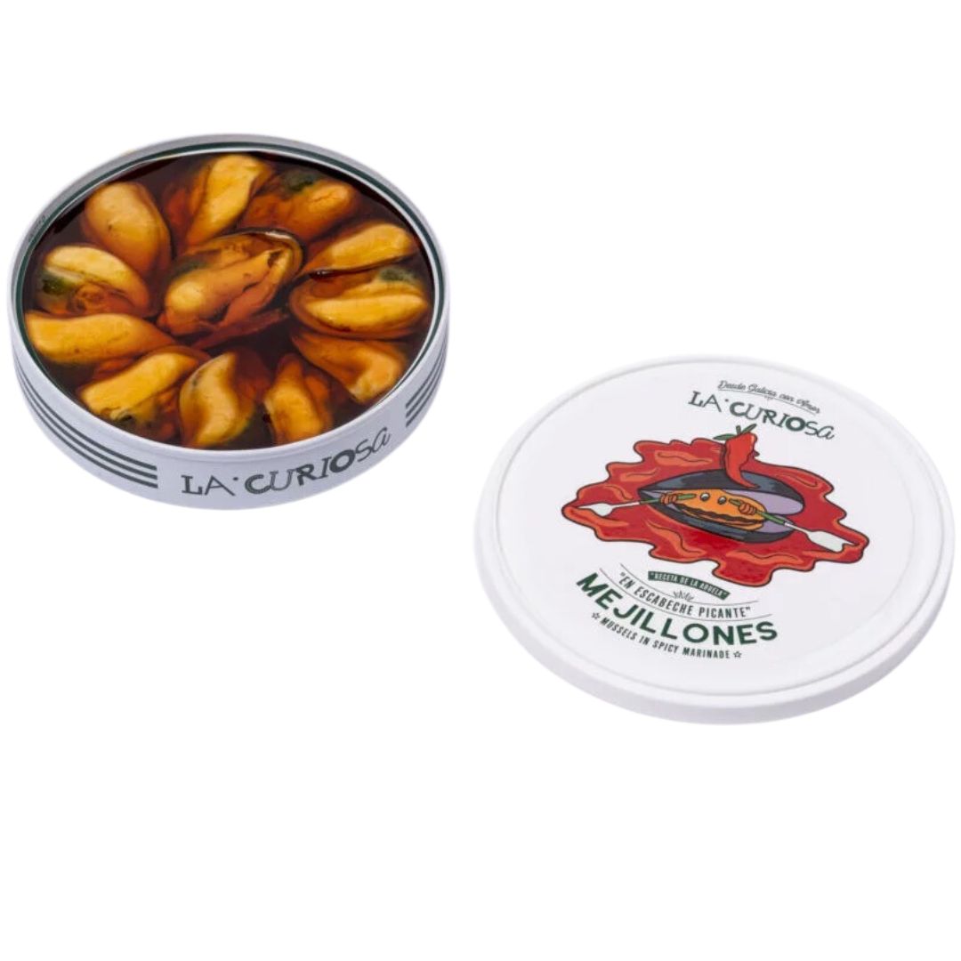 An opened tin of La Curiosa mussels sits beside its playful, illustrated lid. Inside, golden-orange mussels are neatly arranged in a vivid, spicy escabeche marinade, their sheen catching the light. The lid, set aside, shows a bold red splash and a mussel graphic crowned with a chili pepper—an energetic nod to the heat within.