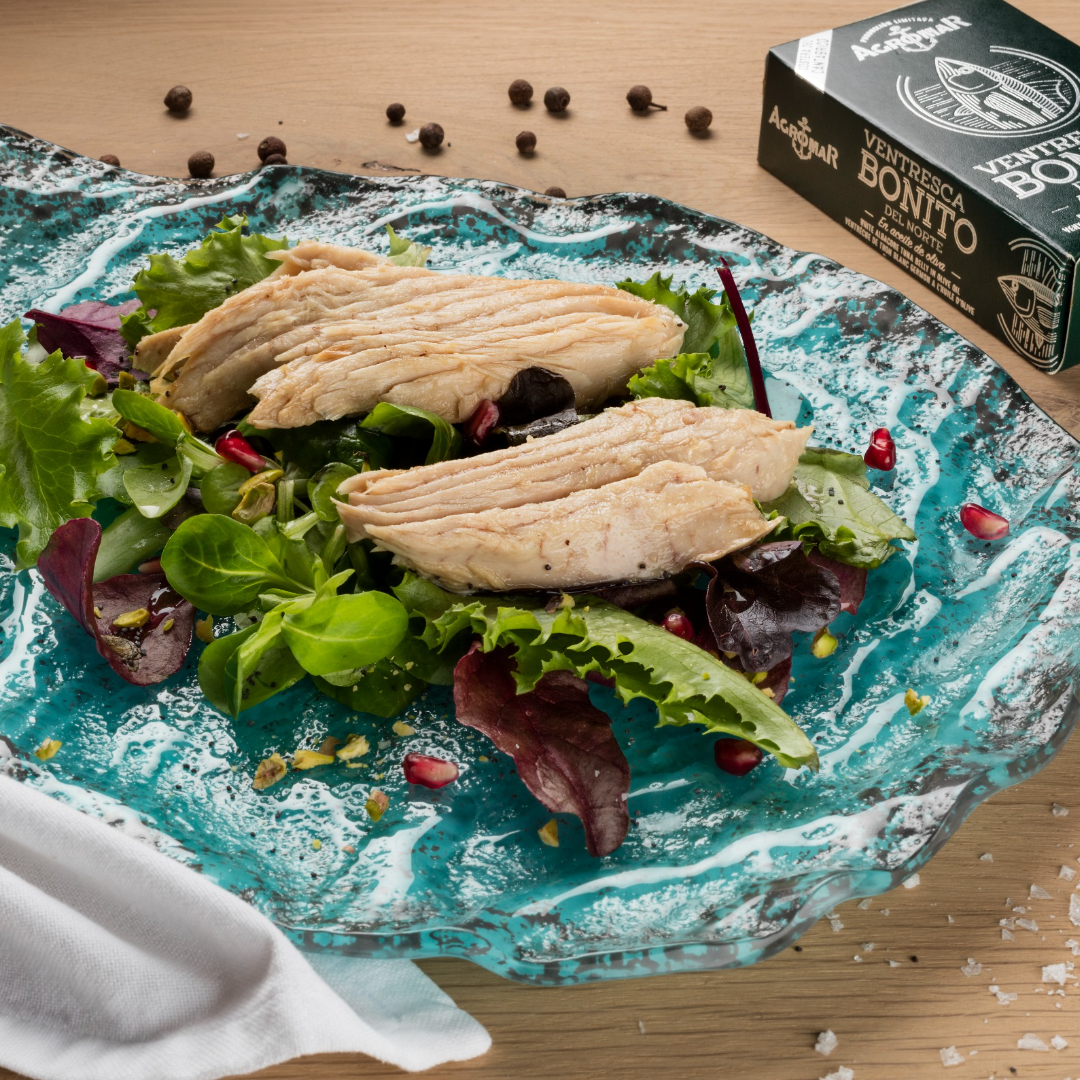 Plate of fresh mixed greens topped with flaky white albacore tuna belly fillets in olive oil. The salad is garnished with pomegranate seeds and sits beside a box of Agromar Ventresca de Bonito del Norte, highlighting premium Spanish tuna from the Cantabrian coast.