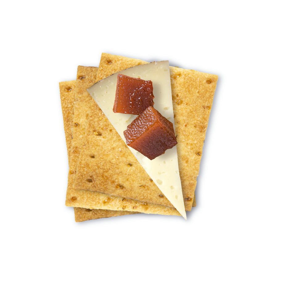A close-up image of three stacked savory olive oil crackers topped with a triangular slice of Manchego cheese and two small cubes of quince paste. The crackers have a golden-brown color and are lightly perforated. The presentation is clean and appetizing, set against a white background.