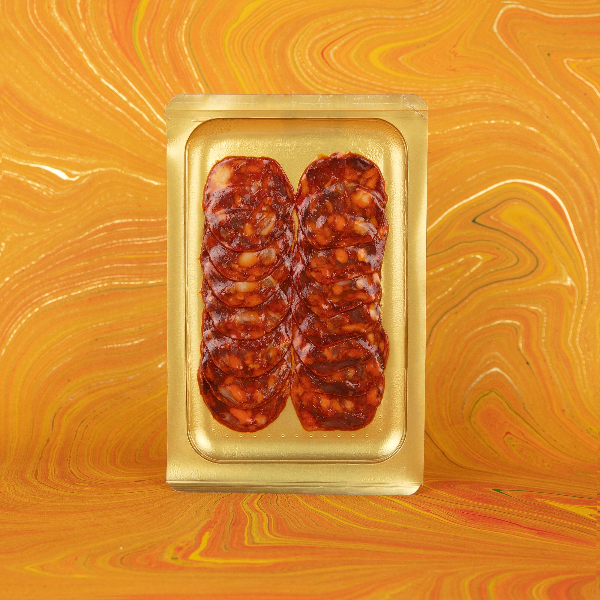 A vibrant display of thinly sliced Iberico Chorizo in a golden vacuum-sealed tray set against a bold orange marbled background. The rich marbling and deep red color of the sausage highlight its premium Spanish origin, ideal for tapas, gourmet platters, and culinary gift selections.