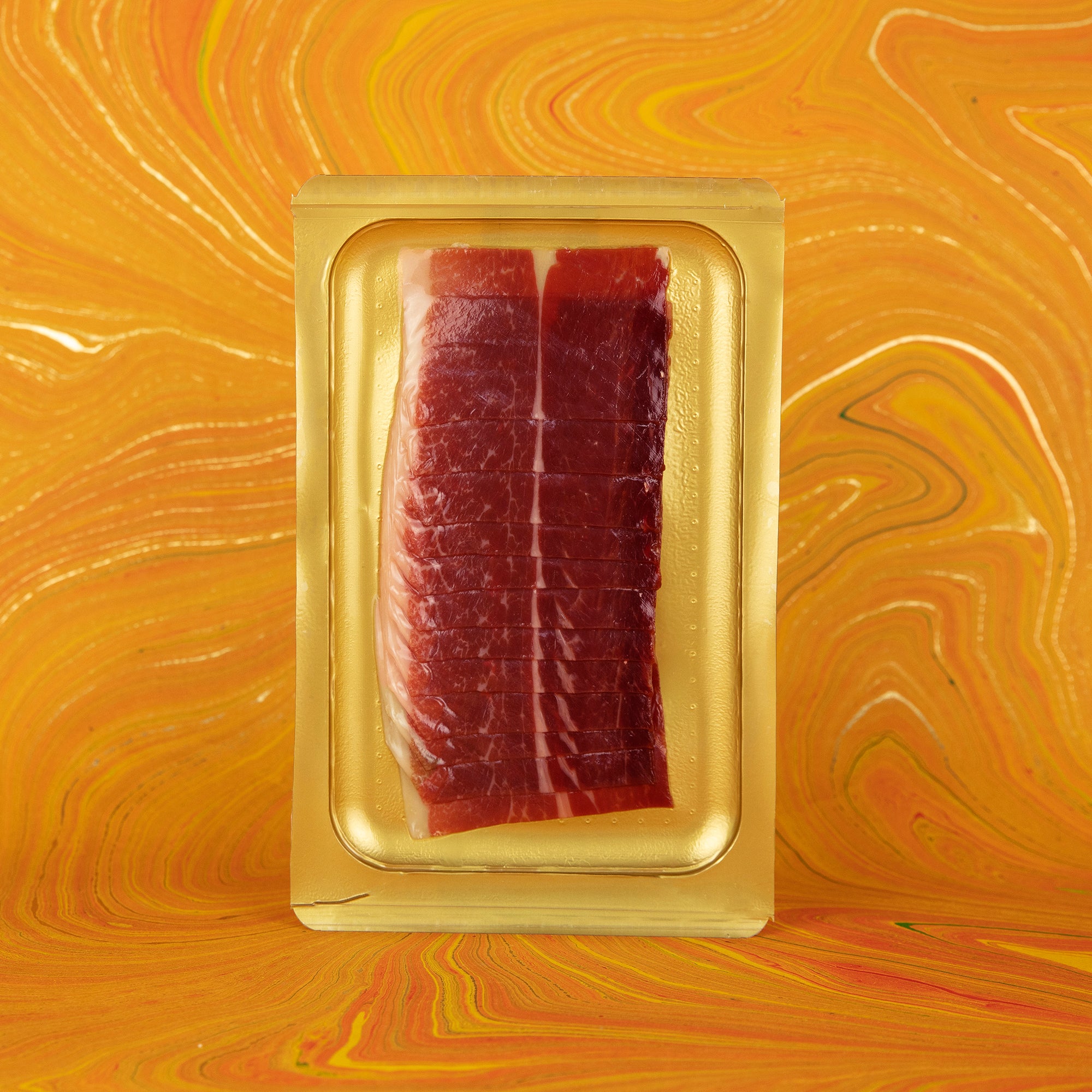 A vacuum-sealed tray of sliced Ibérico ham, presented against a bold orange marbled background. The rich red meat and creamy white fat promise deep flavor and smooth texture.
