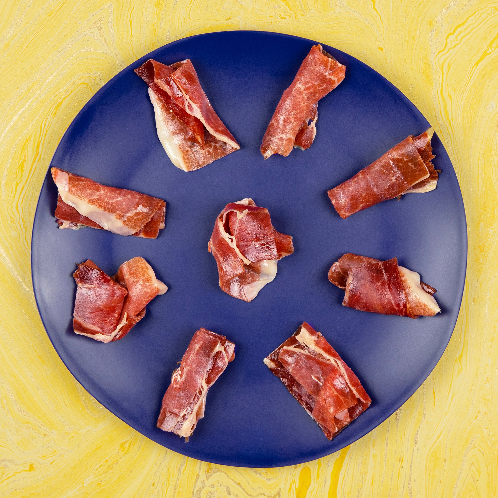 A circular navy-blue plate with neatly arranged pieces of Ibérico ham-wrapped cheese. Each bite-sized tapa is folded with care, making for a savory, elegant appetizer.