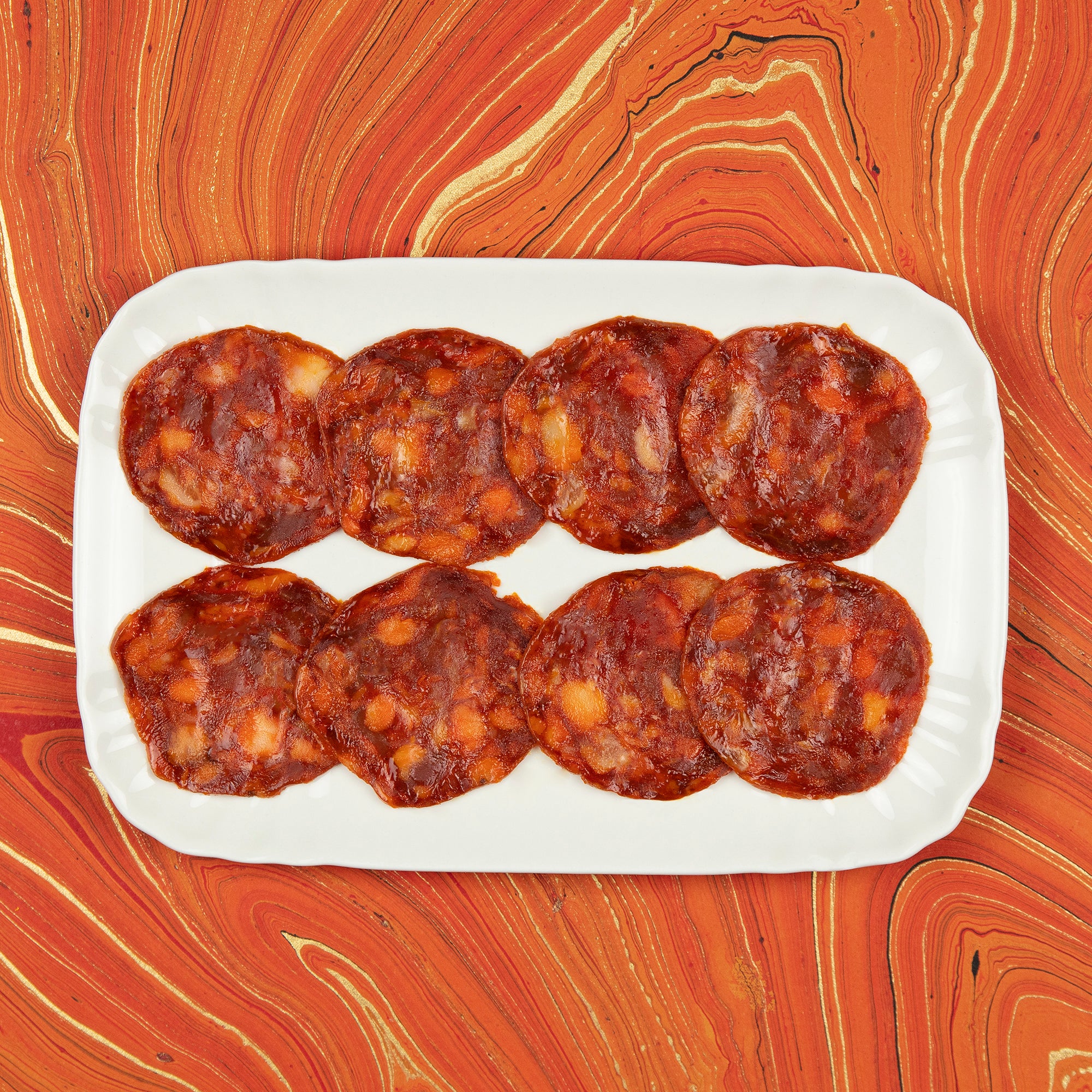 A white rectangular platter neatly arranged with rich, marbled slices of Iberico Chorizo against a vibrant, orange marbled background. The deep red tones and visible fat marbling highlight the premium quality of this traditional Spanish sausage, perfect for gourmet tapas presentations.