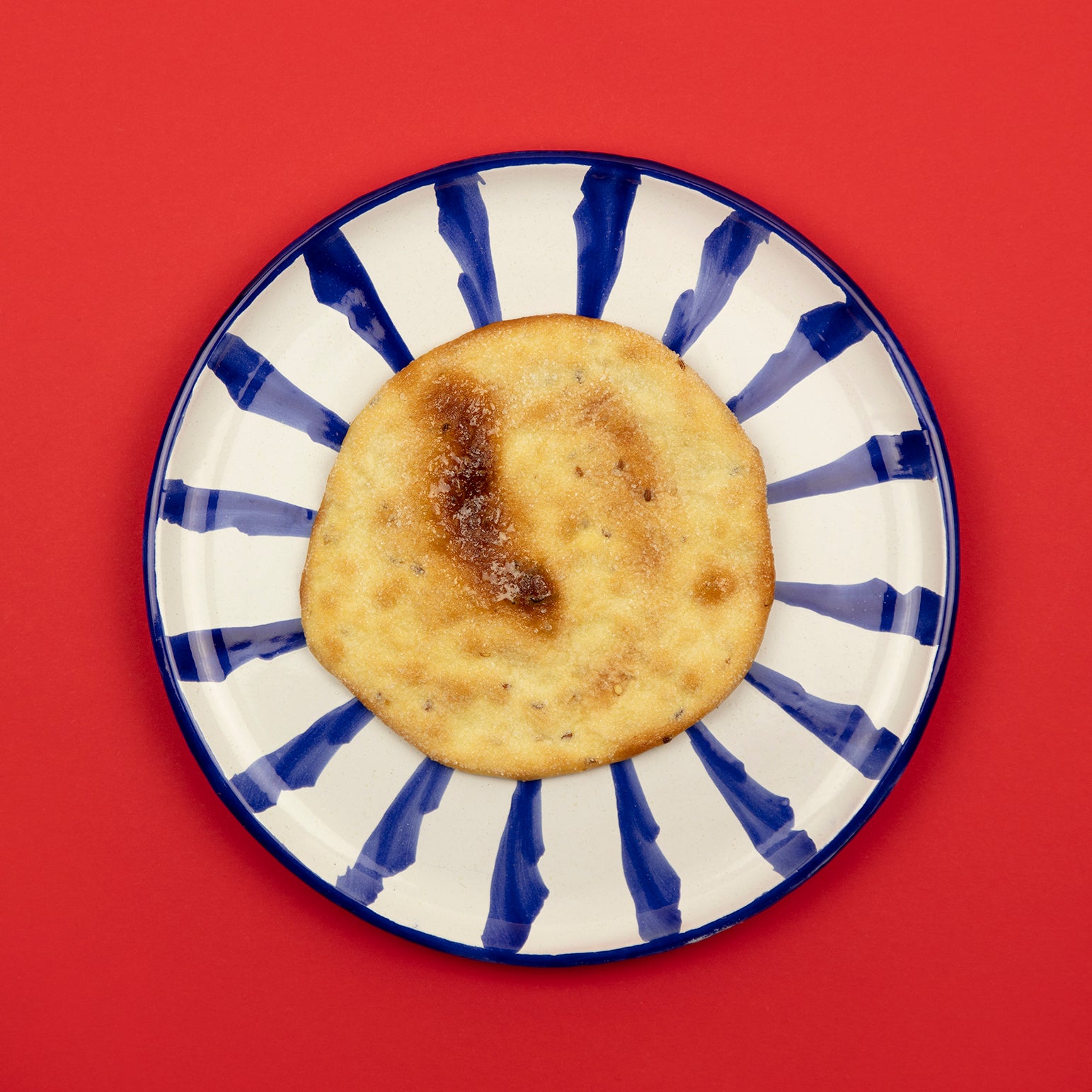 A single Ines Rosales Sweet Olive Oil Torta rests on a bold, hand-painted plate with blue brushstroke rays. Its golden, sugar-speckled surface glows against the vibrant red background, highlighting its artisanal, flaky texture.