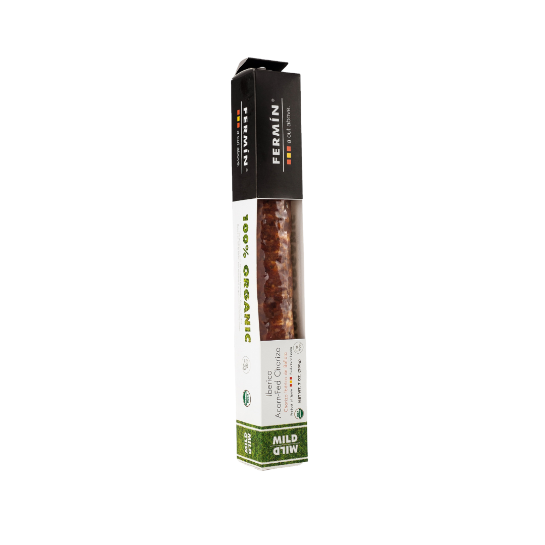 A package of Fermin brand Iberico Acorn-Fed Chorizo sausage, labeled as 100% organic and mild. The packaging features a black top section with the brand name, a white middle section displaying the organic certification and product details, and a green bottom indicating the mild flavor. The chorizo sausage is partially visible through a transparent window in the box.
