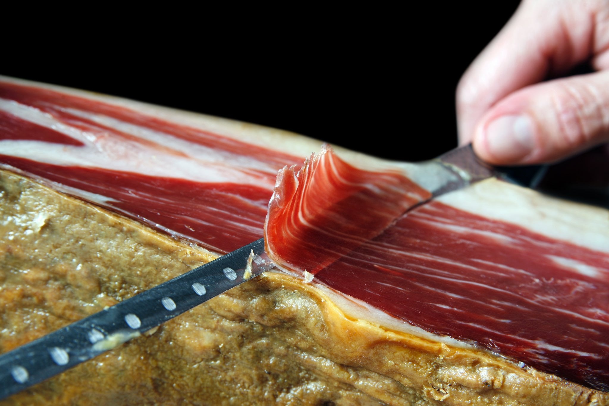 A close-up of a hand slicing a paper-thin piece of Jamón Ibérico from a cured leg. The deep red meat, marbled with silky fat, curls gently as it's lifted—highlighting the craftsmanship behind this prized Spanish delicacy.