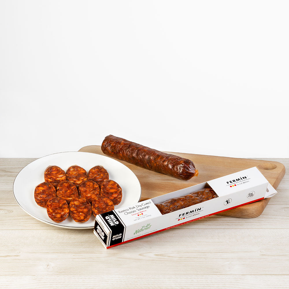 A presentation of Fermin Iberico Pork Dry-Cured Chorizo Sausage. The image includes a full sausage on a wooden cutting board, several chorizo slices arranged on a white plate, and the original product packaging labeled “MILD,” “Naturally,” and “Fermin – a cut above.” The packaging is white with red accents and features a transparent window showing the chorizo inside. The setup is placed on a light wooden surface with a white background.