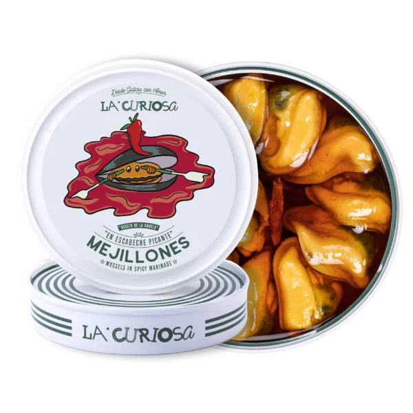 A vibrant tin of La Curiosa mussels bursts open to reveal plump, golden mollusks bathed in fiery escabeche marinade. The lid, whimsically illustrated with a pepper and mussel atop a splash of red, hints at the bold flavors inside. This artisanal conserva from Galicia offers a perfect balance of spice, brine, and tradition.