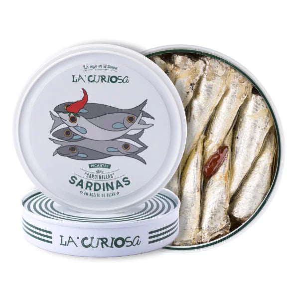 A stylish round tin of La Curiosa spicy sardines in olive oil, featuring a whimsical illustration of sardines stacked with a red chili pepper. The open tin reveals glistening fish fillets, blending artisanal Spanish canning with bold, playful design perfect for gourmet seafood lovers.